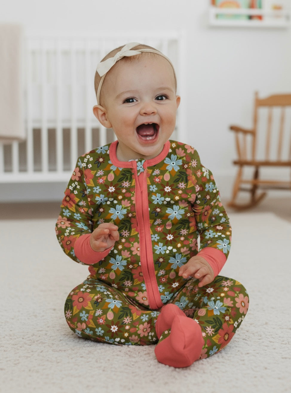 Baby in a floral onesie sitting on a carpeted floor with a crib in the background.