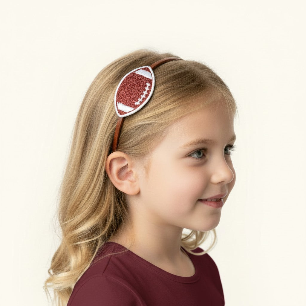 A young girl with blonde hair wearing a brown headband with a football-shaped hair clip, shown in profile against a plain off-white background.