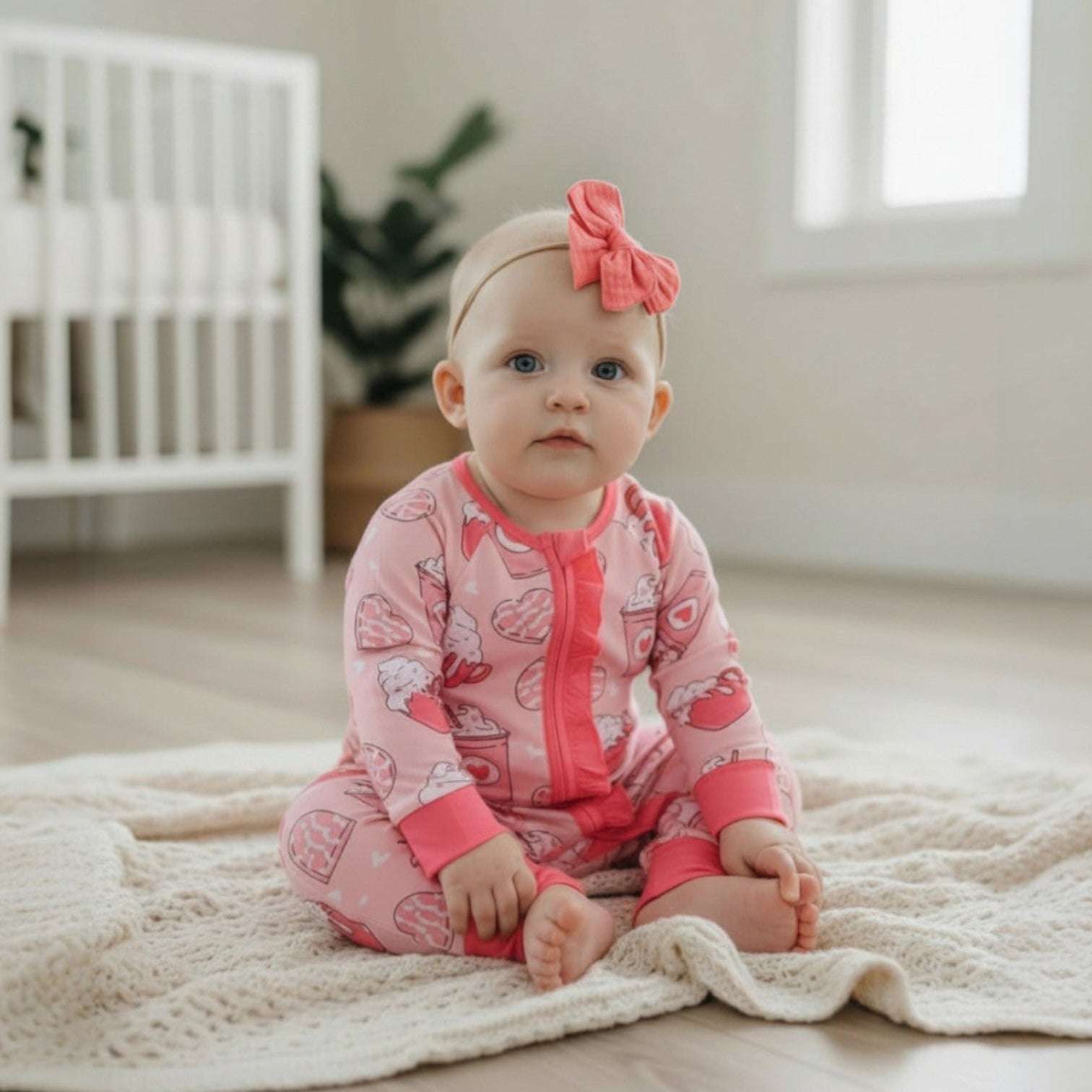 Baby in pink outfit with a bow sitting on a blanket in a nursery.